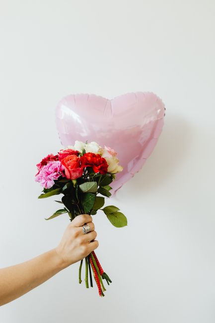 A bouquet of fresh flowers held by a hand, featuring pink, red, and white blossoms with lush green foliage. The arrangement includes roses and hydrangeas, wrapped with a red ribbon around the stems. T