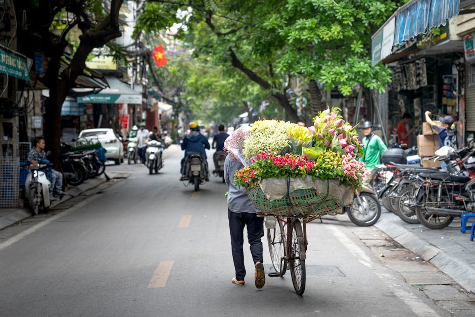 A flower vendor pushes a bicycle loaded with a large, colorful bouquet of fresh flowers, including pink lilies, white hydrangeas, and yellow roses, wrapped in paper and secured with a green basket. Th