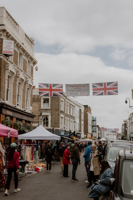 A vibrant street scene on Portobello Road featuring various street stalls and shops beneath Union Jack flags strung across the sky. The foreground shows people browsing and shopping, some engaging in