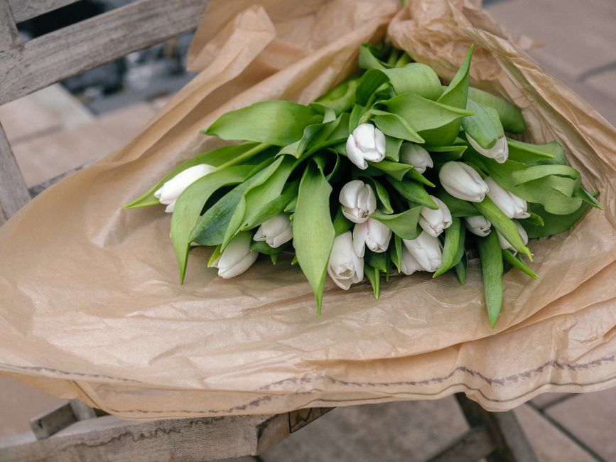 A bouquet of fresh white tulips with green leaves, wrapped in light brown kraft paper, resting on a rustic wooden surface. The tulips are tightly clustered, with petals that appear soft and slightly c