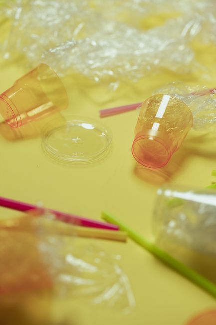 A close-up image features discarded plastic cups, some transparent with a pinkish hue, along with colorful plastic straws in shades of pink, green, and yellow, spread across a bright yellow surface. T