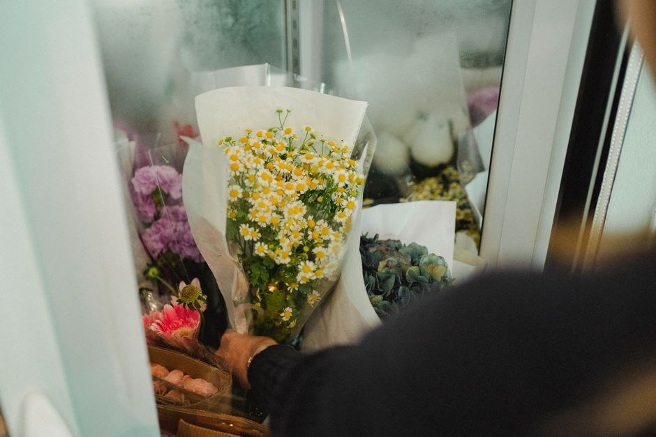 A person holding a bouquet of fresh yellow and white daisies wrapped in white paper, with visible green stems and petals, selecting flowers from a refrigerated display case at floristnottinghill.co.uk