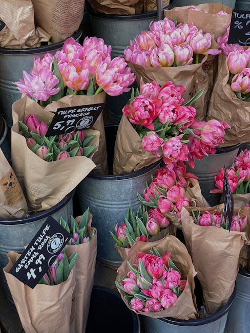 A display of fresh pink tulips with ruffled petals and green leaves, arranged in brown kraft paper wraps and placed inside metal buckets at a floral shop or market stand. Some bouquets are labeled wit
