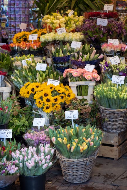 A vibrant display of fresh flowers arranged in baskets and containers at a florist shop, featuring yellow sunflowers, white roses, pink gerbera daisies, purple and pink tulips, and clusters of green t