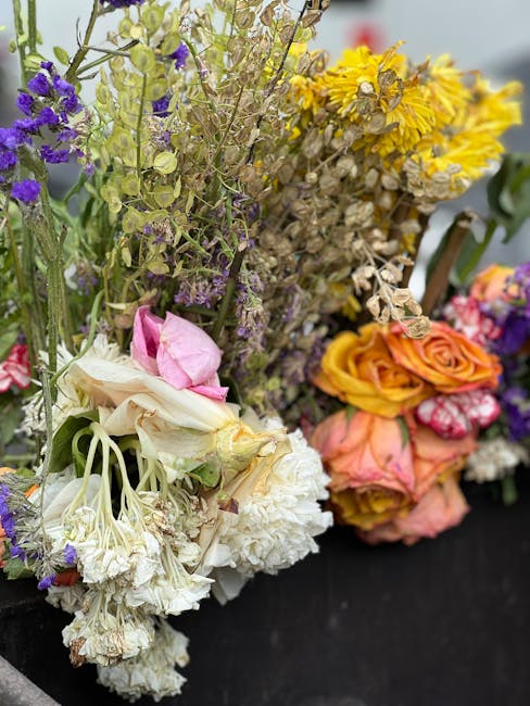 Close-up image of a vibrant floral bouquet featuring a diverse mix of flowers including white carnations, pale pink peonies, purple statice, yellow clusters, and orange roses. The flowers are arranged