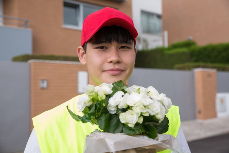 A young delivery person wearing a red cap and a high-visibility yellow vest holds a bouquet of fresh white roses with lush green leaves, wrapped in white paper. The bouquet is positioned in front of t