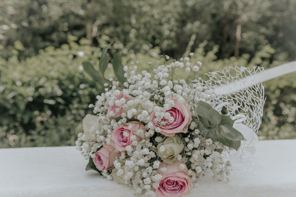 A delicate floral bouquet featuring soft pink roses and white ranunculus, complemented by sprigs of baby's breath and lush green foliage. The arrangement is wrapped in a white netted lace material, wi