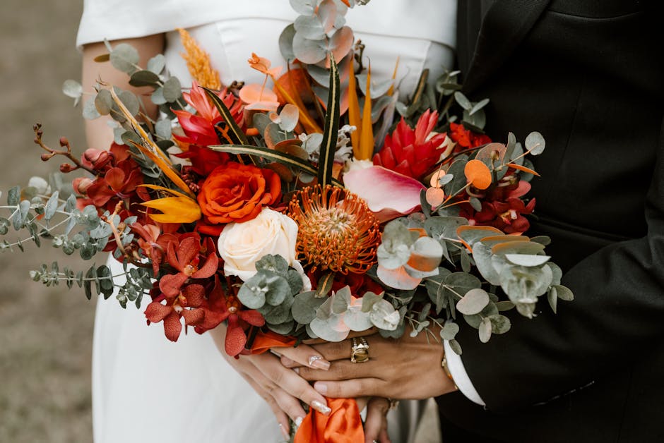 A floral arrangement featuring a vibrant bouquet held by a bride and groom, comprising orange and red roses, pink and white calla lilies, a protea flower, and sprigs of eucalyptus with round, silvery-